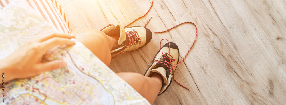Young female inspecting a city map resting on the bed with untied trekking boots on wooden floor - obrazy, fototapety, plakaty 