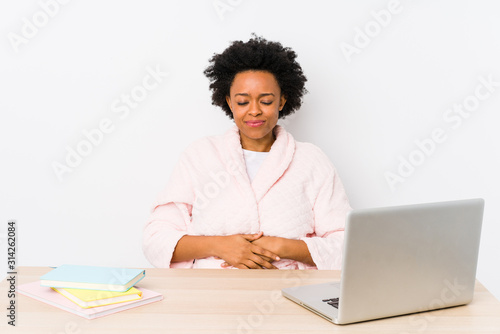 Middle aged african american woman working at home isolated touches tummy, smiles gently, eating and satisfaction concept.