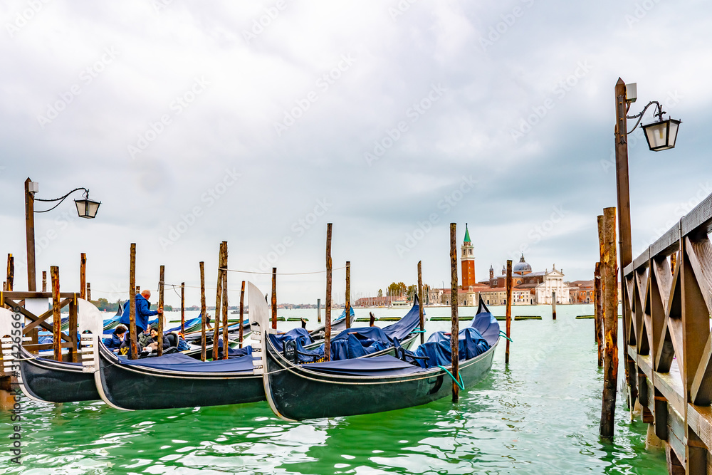 Venice, Italy. Gondolas docked by wooden mooring poles. Tourist passengers/ people enter for famous romantic tour ride. San Giorgio Maggiore Basilica Church Bell Tower in background