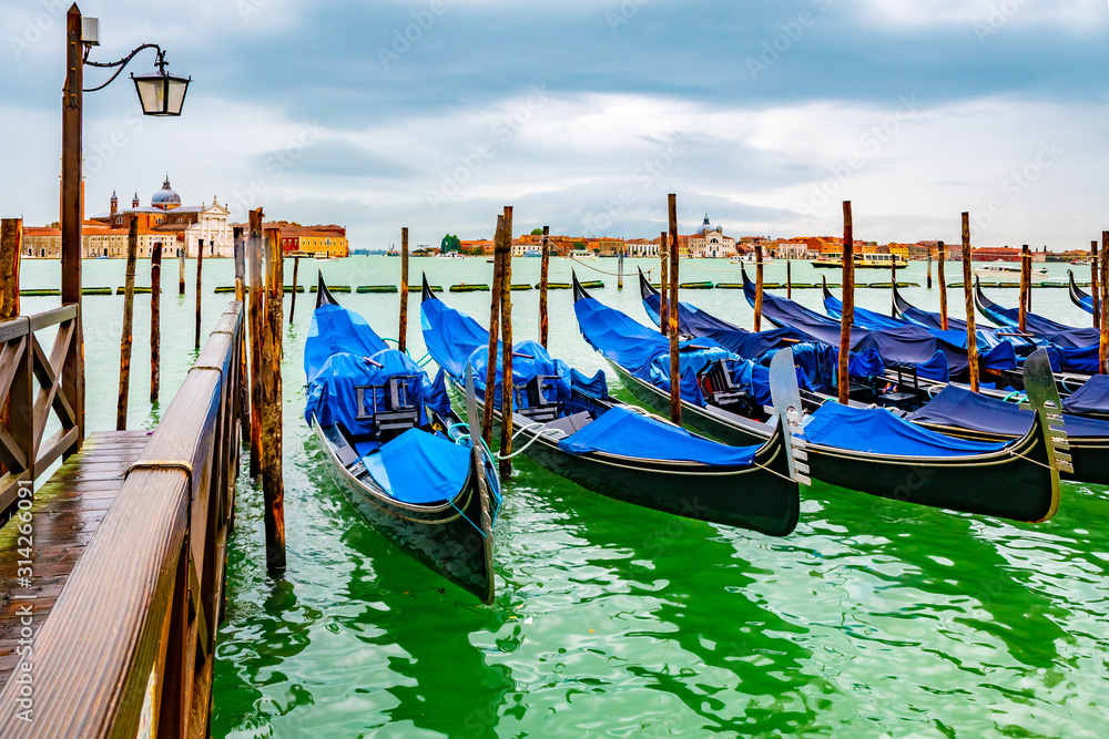 Venice, Italy. Empty gondolas docked on lagoon canal channel coast by wooden mooring poles and dock boardwalk. Famous romantic tourist boat ride experience. San Giorgio Maggiore Basilica church behind