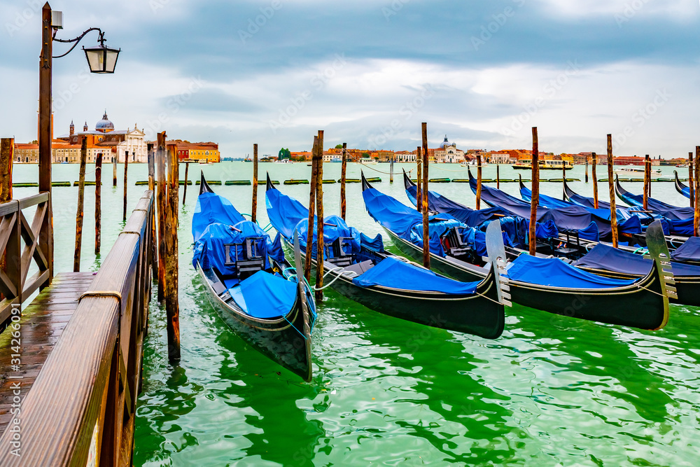 Fototapeta premium Venice, Italy. Empty gondolas docked on lagoon canal channel coast by wooden mooring poles and dock boardwalk. Famous romantic tourist boat ride experience. San Giorgio Maggiore Basilica church behind