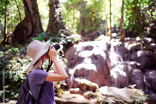 Travel and technology. Young woman in hat with rucksack taking photo of waterfall in jungle.