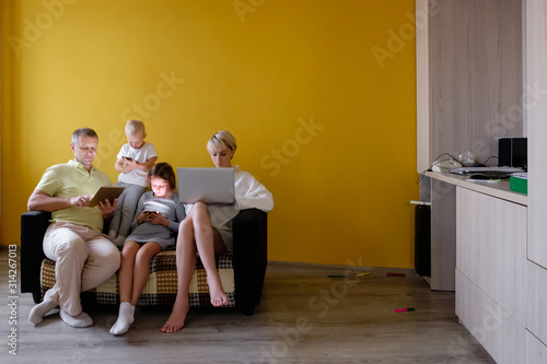 Side view of happy big family dad mom daughter and son are watching gadgets using high speed internet while sitting on sofa on yellow background. Internet addiction concept