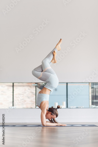 Strong young girl yoga instructor doing vrischikasana in white hall on the floor. The concept of long workouts willpower and endurance. Copyspace. Pincha Mayurasana exercise, handstand pose