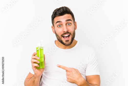 Young caucasian man holding a moisturizer with aloe vera isolated smiling cheerfully pointing with forefinger away.