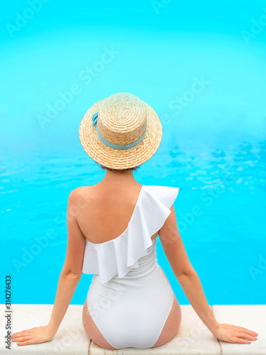 beautiful redhead (ginger) woman's back in the white swimminig suit sitting by the swimming pool (or sea or ocean) in the straw hat in summer day. Summer, relax, wellness, recreation concept