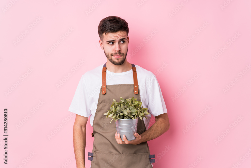 Young caucasian gardener man holding a plant isolated confused, feels doubtful and unsure. Young caucasian gardener man holding a plant isolated confused, feels doubtful and unsure.