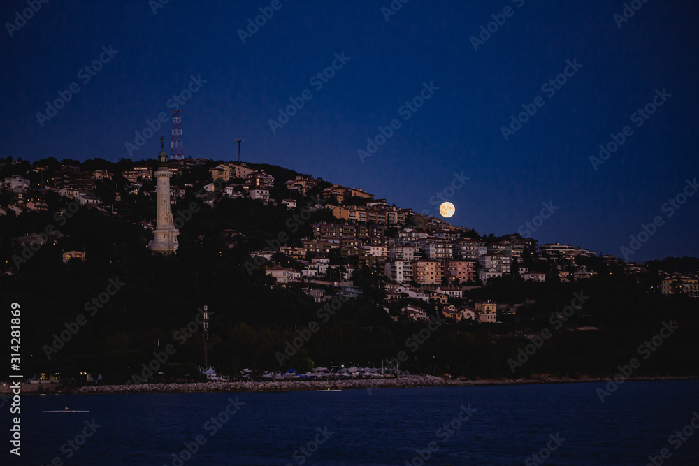 Trieste city skyline view at night with full moon rising from the mediterranean Sea. Famous light tower visible.