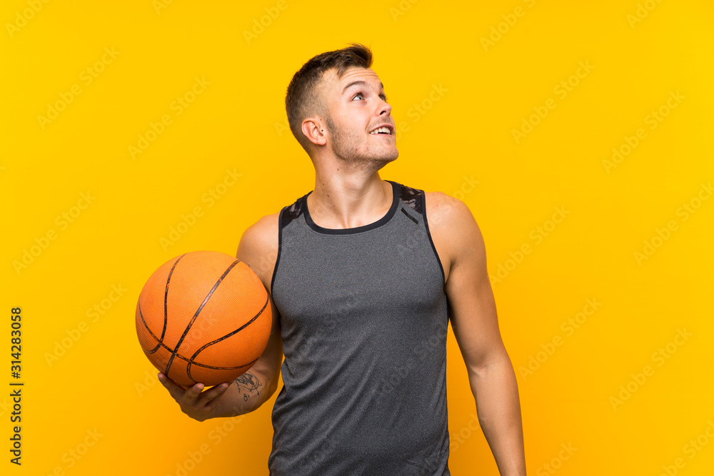 Young handsome blonde man holding a basket ball over isolated yellow background looking up while smiling