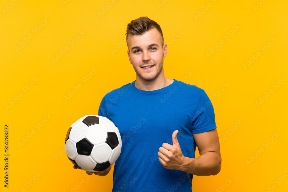 Young handsome blonde man holding a soccer ball over isolated yellow background with thumbs up because something good has happened