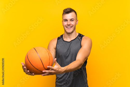 Happy Young handsome blonde man holding a basket ball over isolated yellow background