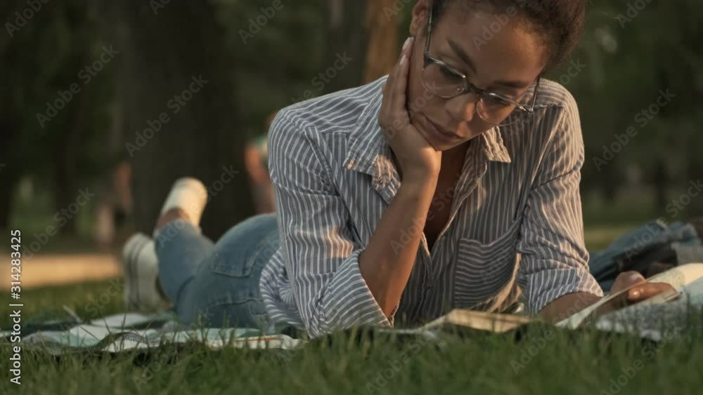 Concentrated pretty african woman in eyeglasses reading book while lying on plaid in the park Concentrated pretty african woman in eyeglasses reading book while lying on plaid in the park