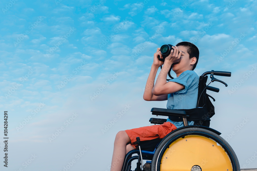 Asian special child on wheelchair is happily on blue sky and cloud background,Excited to travel on a holiday, He has camera, Life in the education age of disabled children, Happy disabled kid concept.