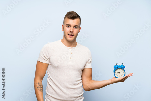 Young handsome blonde man over isolated blue background holding vintage alarm clock