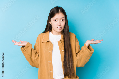 Obraz na plátně Young chinese woman posing in a blue background isolated confused and doubtful shrugging shoulders to hold a copy space