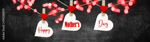 Happy Mother's Day background banner - White hearts hang on wooden clothes pegs with wooden hearts on a string isolated on rustic black concrete stone texture and bokeh lights, with space for text
