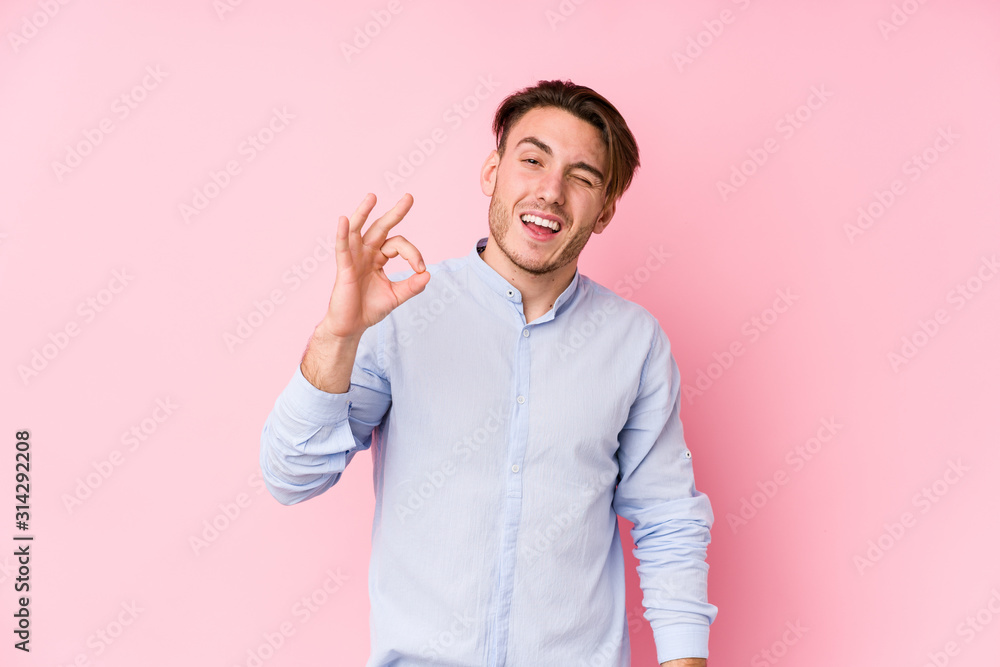Young caucasian man posing in a pink background isolated winks an eye and holds an okay gesture with hand. Young caucasian man posing in a pink background isolated winks an eye and holds an okay gesture with hand.