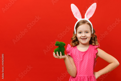 Happy easter. Cute little girl in bunny ears holds a green rabbit. Closeup baby on a red colored background