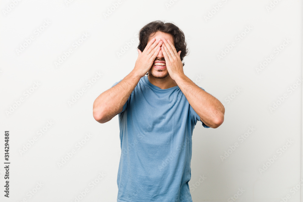 Young handsome man against a white background covers eyes with hands, smiles broadly waiting for a surprise. Young handsome man against a white background covers eyes with hands, smiles broadly waiting for a surprise.
