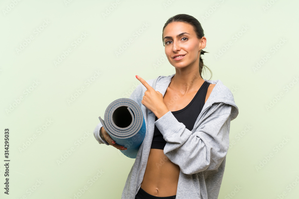 Young sport woman with mat over isolated green background pointing to the side to present a product Young sport woman with mat over isolated green background pointing to the side to present a product
