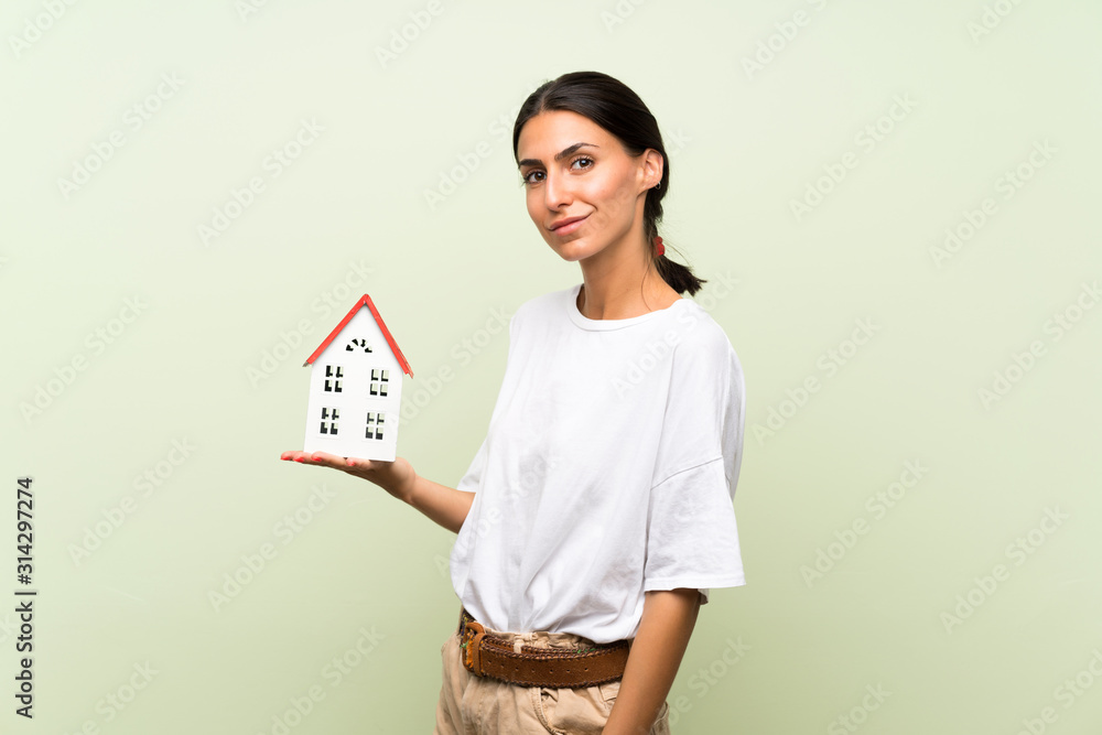 Young woman over isolated green background holding a little house Young woman over isolated green background holding a little house