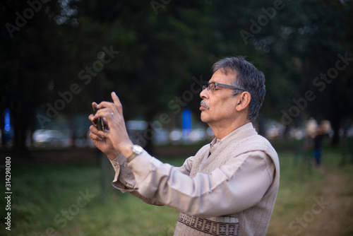 Side portrait of an old Indian Bengali man in woolen sweater taking photograph of sky and trees by his cellphone in a field in winter afternoon in green background. Indian lifestyle and winter.