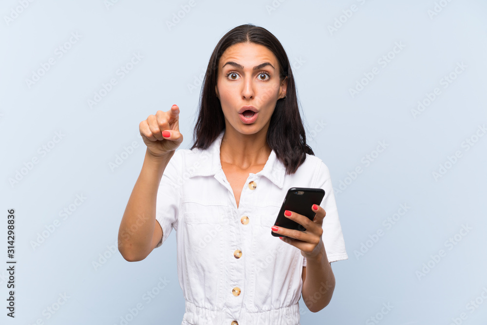 Young woman with a mobile phone over isolated blue wall surprised and pointing front Young woman with a mobile phone over isolated blue wall surprised and pointing front