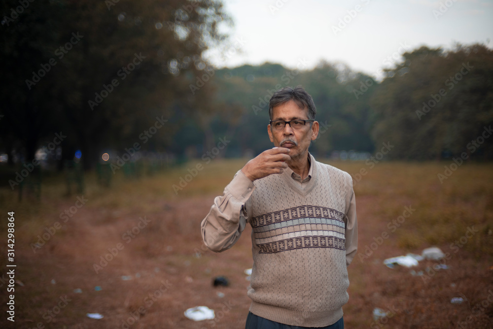 Portrait of an old Indian Bengali man in woolen sweater having tea/coffee in a field in winter afternoon in natural background. Indian lifestyle and winter. Portrait of an old Indian Bengali man in woolen sweater having tea/coffee in a field in winter afternoon in natural background. Indian lifestyle and winter.