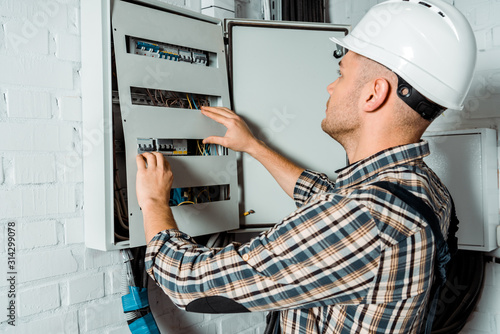 electrician in safety helmet looking at switchboard near brick wall