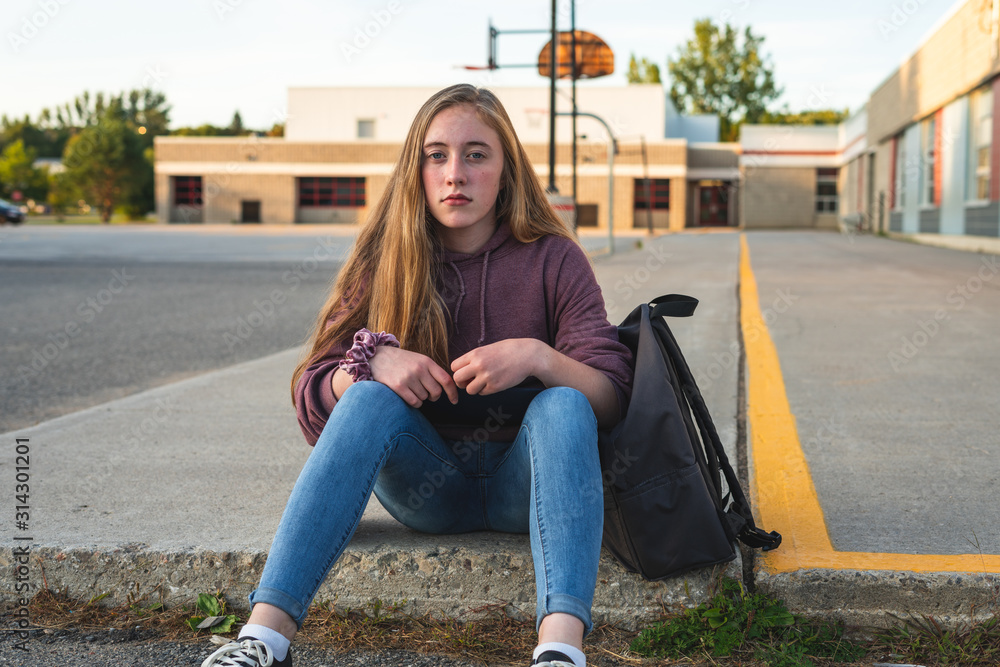 Depressed/Sad teen girl sitting on a curb in front of a high school during sunset while sitting next to a backpack, holding a binder.