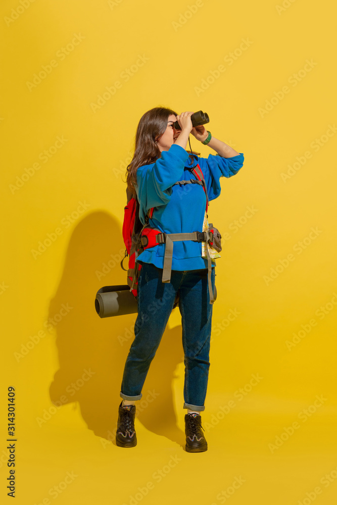 Smiling, looking up. Portrait of a cheerful young caucasian tourist girl with bag and binoculars isolated on yellow studio background. Preparing for traveling. Resort, human emotions, vacation. Smiling, looking up. Portrait of a cheerful young caucasian tourist girl with bag and binoculars isolated on yellow studio background. Preparing for traveling. Resort, human emotions, vacation.