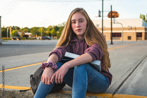 Depressed/Sad teen girl sitting on a curb in front of a high school during sunset while sitting next to a backpack, holding binders and a smartphone.