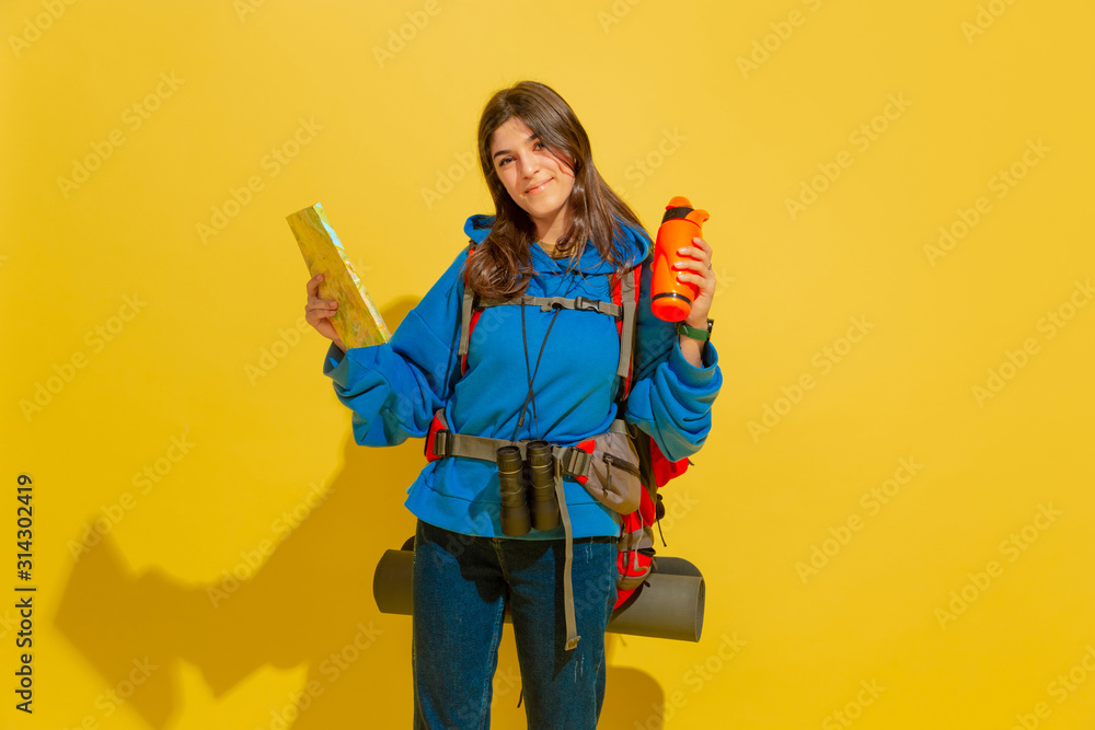 Smiling, looks dreamful. Portrait of a cheerful young caucasian tourist girl with bag and binoculars isolated on yellow studio background. Preparing for traveling. Resort, human emotions, vacation. Smiling, looks dreamful. Portrait of a cheerful young caucasian tourist girl with bag and binoculars isolated on yellow studio background. Preparing for traveling. Resort, human emotions, vacation.