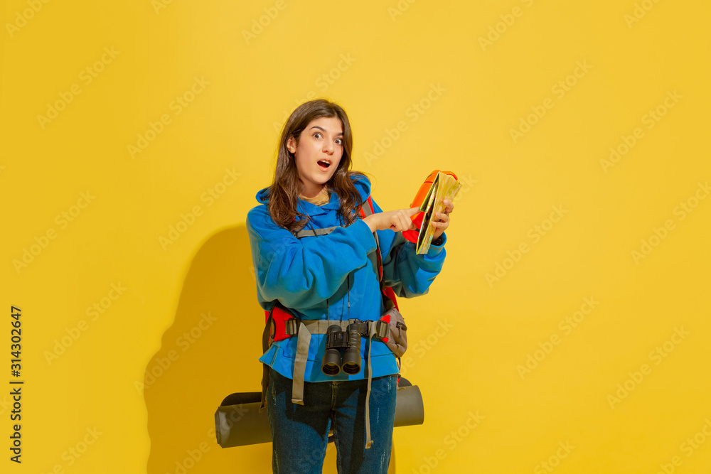 Smiling, pointing on map. Portrait of a cheerful young caucasian tourist girl with bag and binoculars isolated on yellow studio background. Preparing for traveling. Resort, human emotions, vacation. Smiling, pointing on map. Portrait of a cheerful young caucasian tourist girl with bag and binoculars isolated on yellow studio background. Preparing for traveling. Resort, human emotions, vacation.