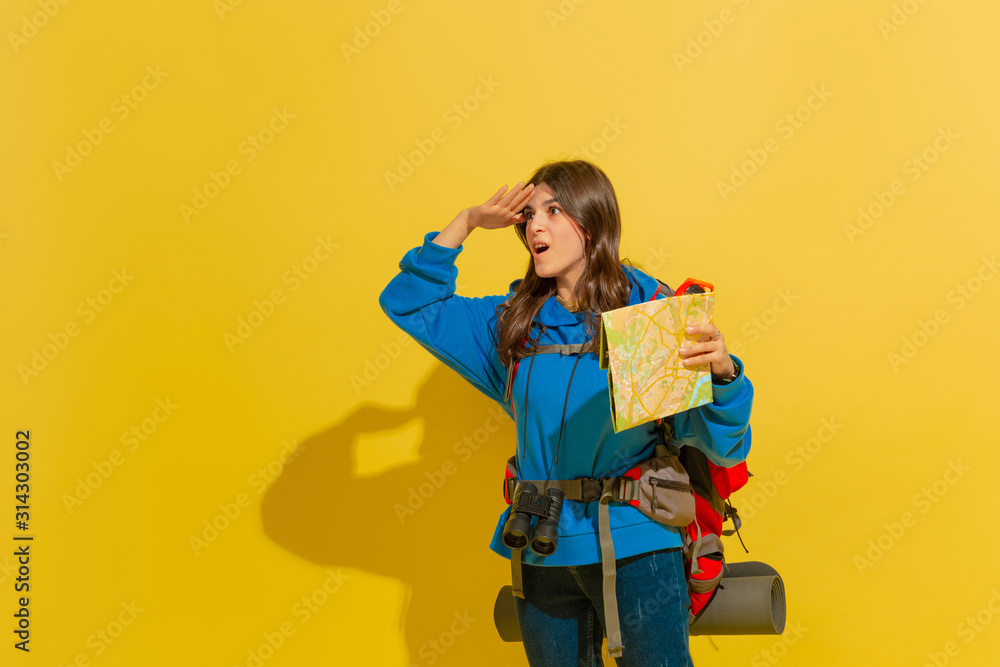 Smiling, looking for way. Portrait of a cheerful young caucasian tourist girl with bag and binoculars isolated on yellow studio background. Preparing for traveling. Resort, human emotions, vacation. Smiling, looking for way. Portrait of a cheerful young caucasian tourist girl with bag and binoculars isolated on yellow studio background. Preparing for traveling. Resort, human emotions, vacation.