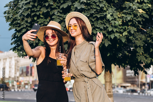 Two pretty girls take selfie in the summer outdoors at sunset. Girlfriends have fun, laugh, smile and take pictures. Closeup portrait of two young women in straw hats.