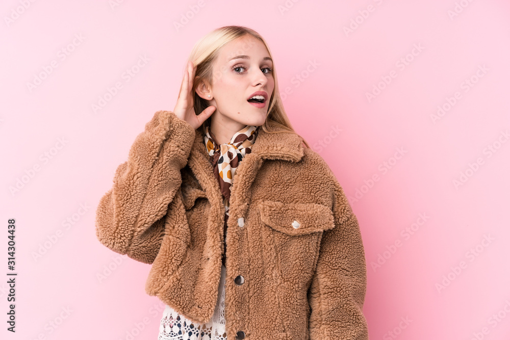 Young blonde woman wearing a coat against a pink background trying to listening a gossip. Young blonde woman wearing a coat against a pink background trying to listening a gossip.