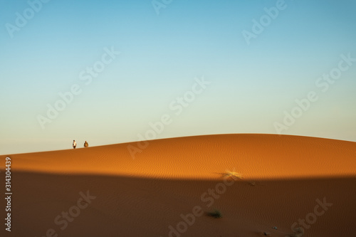 Fototapeta Naklejka Na Ścianę i Meble -  Dos personas caminando sobre dunas en el desierto al atardecer. Erg Chebbi, Marruecos.