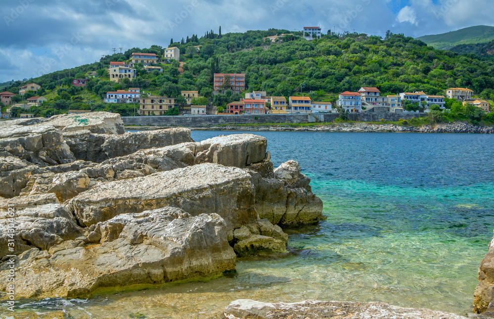 Beautiful summer landscape of the resort with blue sea water, cliffs and rocks on the shore, village with colorful houses and mountains on the horizon. Corfu Island, Greece. 