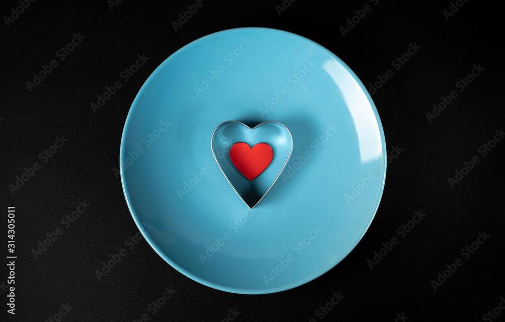 Fototapeta premium Red heart in a metal shape on a blue plate on a black background, concept for Valentine's Day, wedding, birthday and other holidays, view from the top.