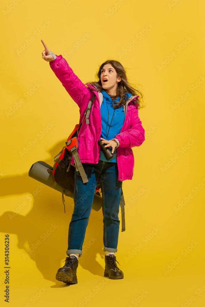 Astonished pointing up. Portrait of a cheerful young caucasian tourist girl with bag and binoculars isolated on yellow studio background. Preparing for traveling. Resort, human emotions, vacation. Astonished pointing up. Portrait of a cheerful young caucasian tourist girl with bag and binoculars isolated on yellow studio background. Preparing for traveling. Resort, human emotions, vacation.