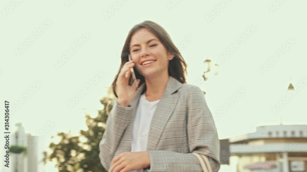 A good-looking smiling girl is having a phone call while staying outside in the city center in summer A good-looking smiling girl is having a phone call while staying outside in the city center in summer