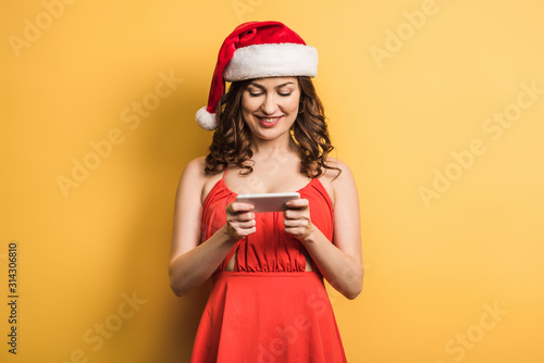 cheerful girl in santa hat messaging on smartphone on yellow background