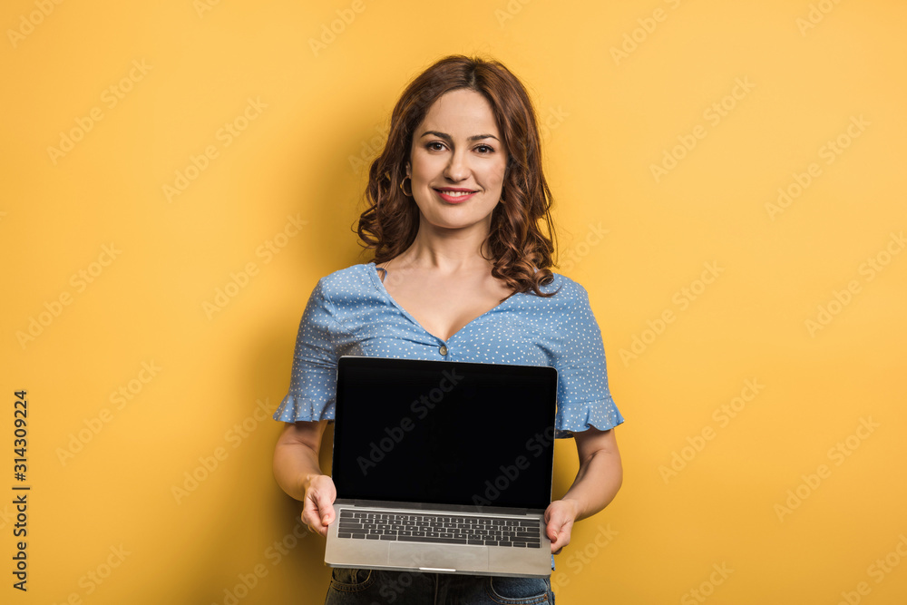 Naklejka premium smiling woman holding laptop with blank screen on yellow background