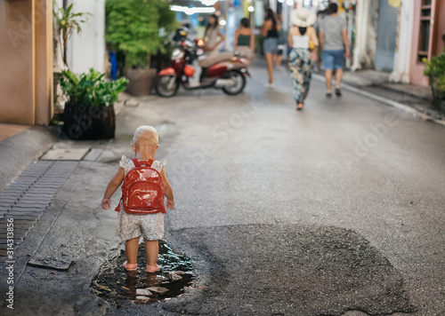 A cute child with a red backpack stands in a puddle with his back along the middle of the street in the evening.