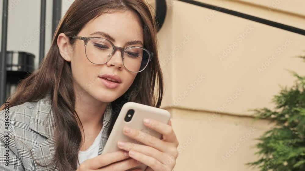 An attractive girl wearing glasses is chatting with someone using her smartphone while sitting outside in the city center An attractive girl wearing glasses is chatting with someone using her smartphone while sitting outside in the city center
