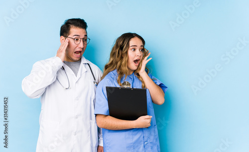 Young doctor couple posing in a blue background isolated shouts loud, keeps eyes opened and hands tense.