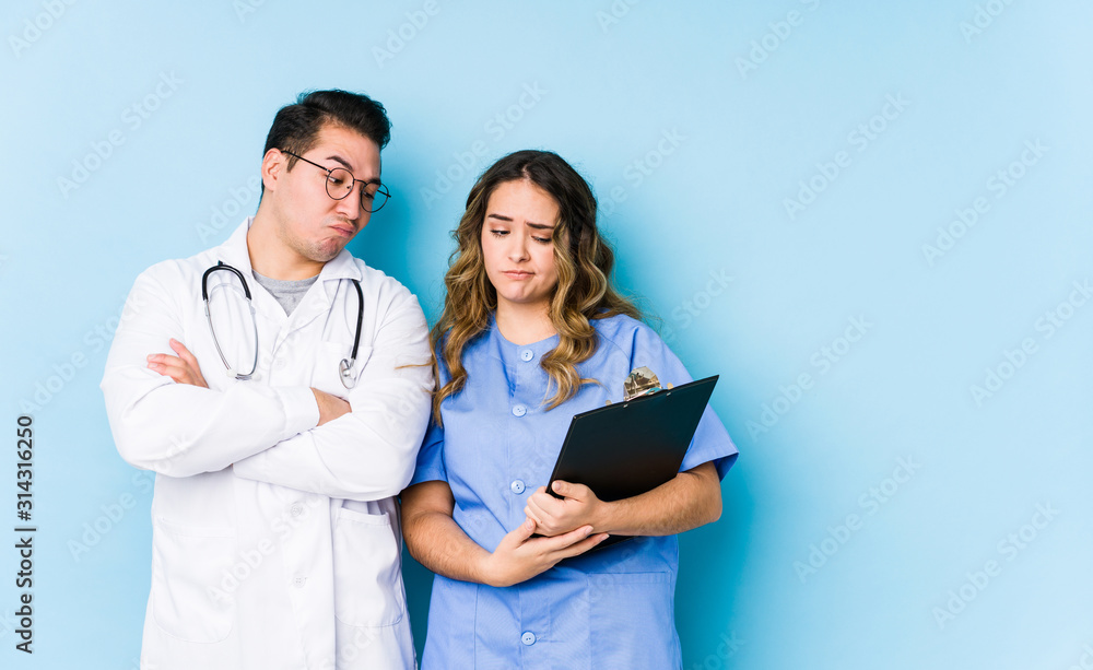 Young doctor couple posing in a blue background isolated unhappy looking in camera with sarcastic expression. Young doctor couple posing in a blue background isolated unhappy looking in camera with sarcastic expression.