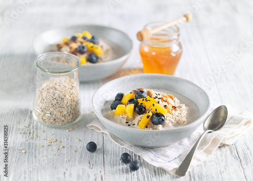 Oatmeal porridge with mango, blueberries, and chopped almonds on white wooden table. Jar of honey on background. Selective focus. Bright healthy breakfast image on a light background, space for text.