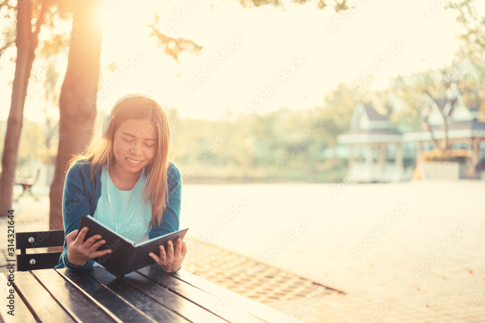 Smiling woman in eyeglasses reading book and listening to music with headphones in park outdoor against sunlight,City lifestyle concept.